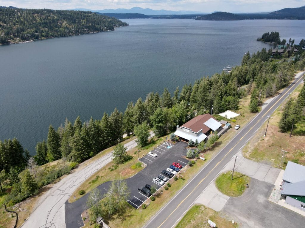 Aerial drone view of a freshly repaved commercial parking lot adjacent to a two-lane highway, surrounded by evergreen trees along the shoreline of a large lake in North Idaho.