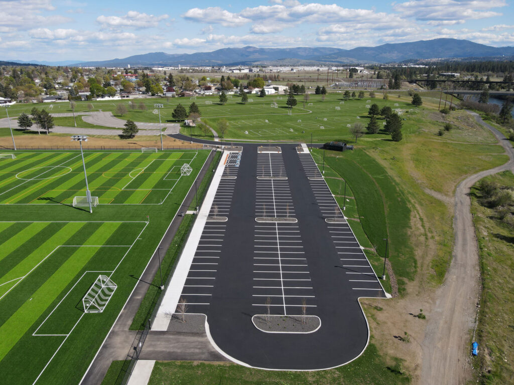 Aerial drone photograph of a freshly paved parking lot adjacent to multiple synthetic turf soccer fields, featuring crisp white striping, ADA stalls, and curbed landscape islands.
