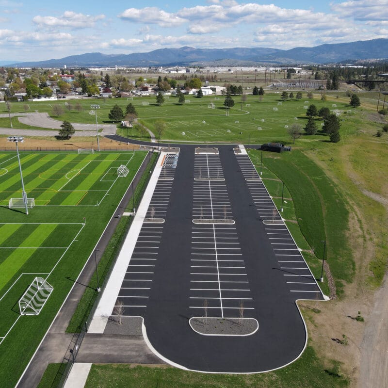 Aerial drone photograph of a freshly paved parking lot adjacent to multiple synthetic turf soccer fields, featuring crisp white striping, ADA stalls, and curbed landscape islands.