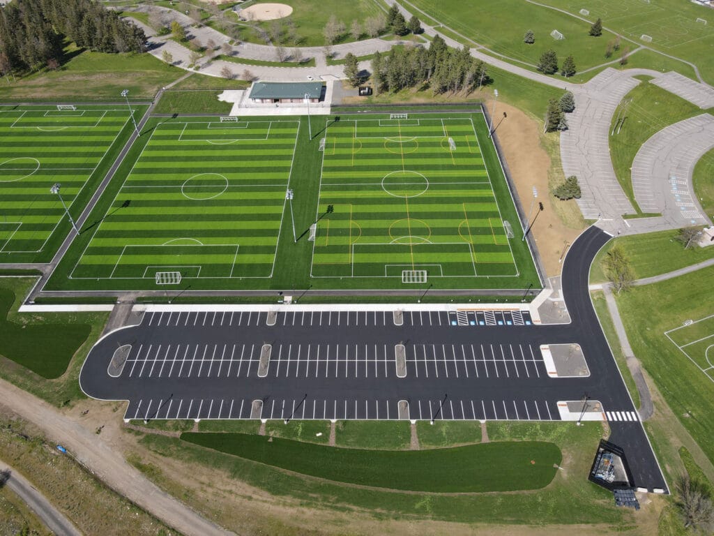 Drone Perspective-Sports Complex Parking, Spokane Valley-WA Oblique aerial drone photo of a newly paved sports complex parking lot with white line striping, curbed islands with young trees, and synthetic turf soccer fields in the foreground, with the Spokane River valley and Selkirk Mountains visible in the background.
