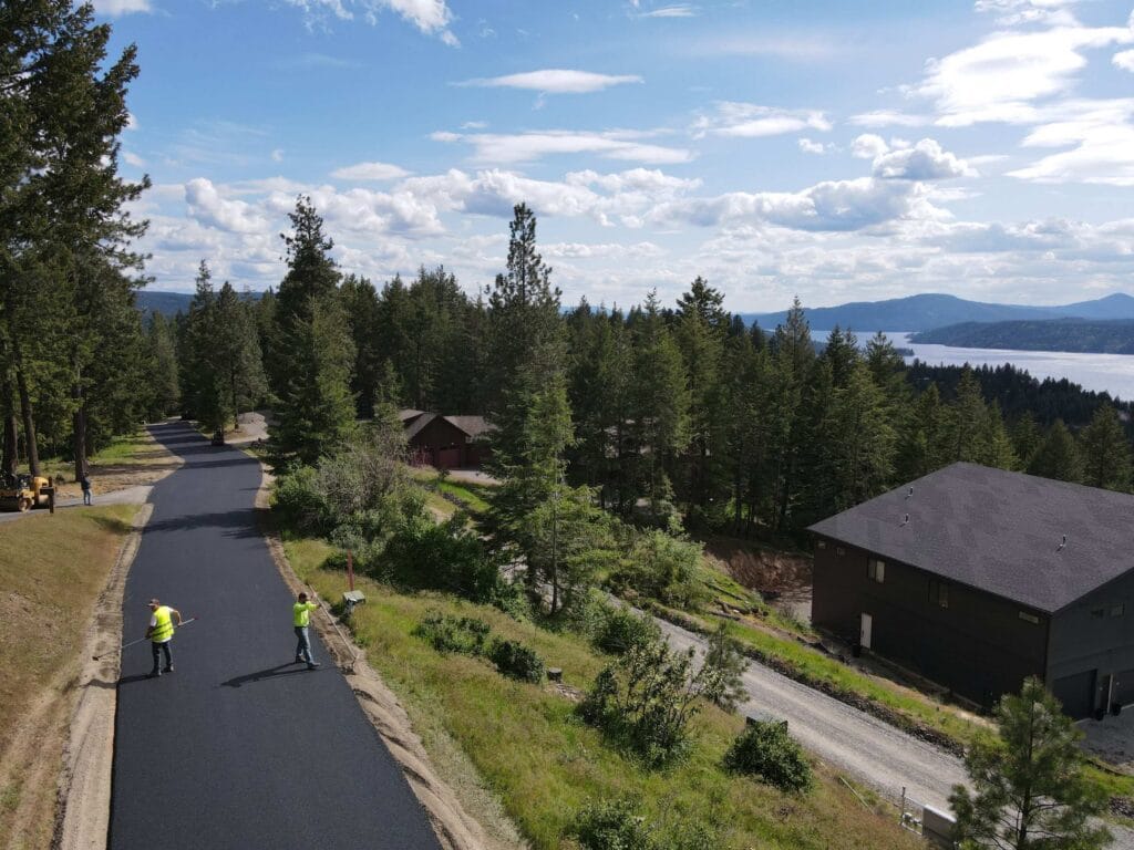 Aerial view of Asphalt Pros USA crew finishing a newly paved private road surrounded by forested residential properties in Coeur d'Alene, Idaho.